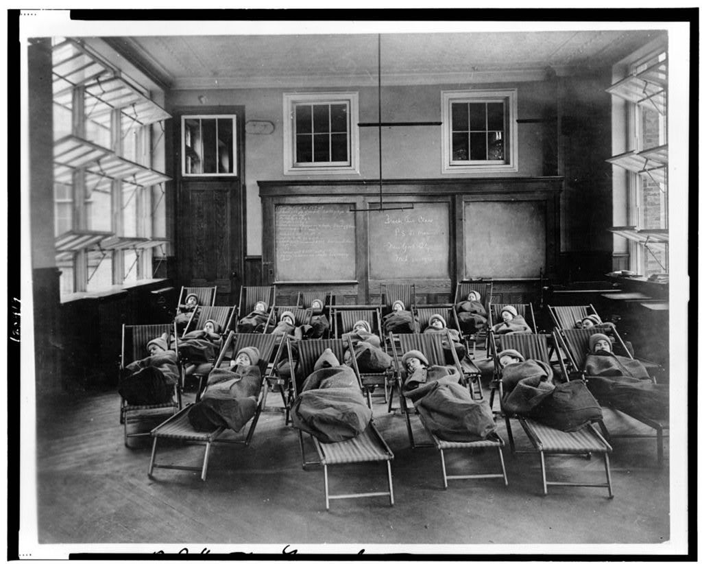 Children in a fresh air class with open windows on cots and wrapped in blankets in 1911.